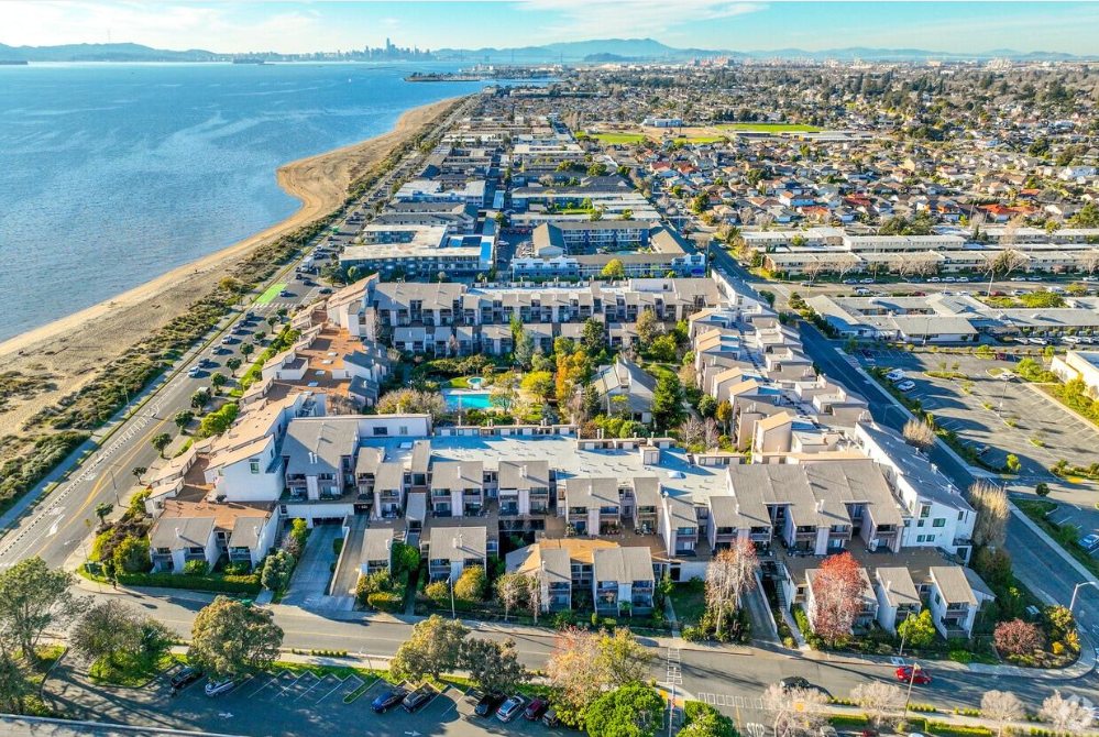 Aerial view of Balena Village: multi-story apartment buildings arranged around landscaped courtyards and a pool, beside a beach and the bay, with a city skyline in the distance.