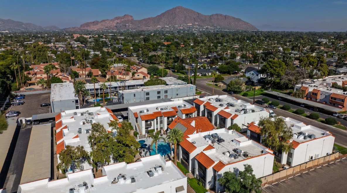 Aerial view of Villa Toscana: white buildings with red tile roofs arranged around a central courtyard pool, with palm trees and mountains in the background.